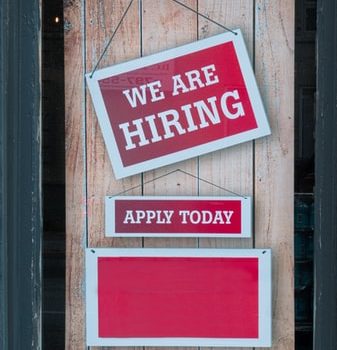 Bewerbungsfrist als Koordinations- und Verwaltungskraft verlängert Rotes Schild an Holztür, auf dem "We are hiring" steht.