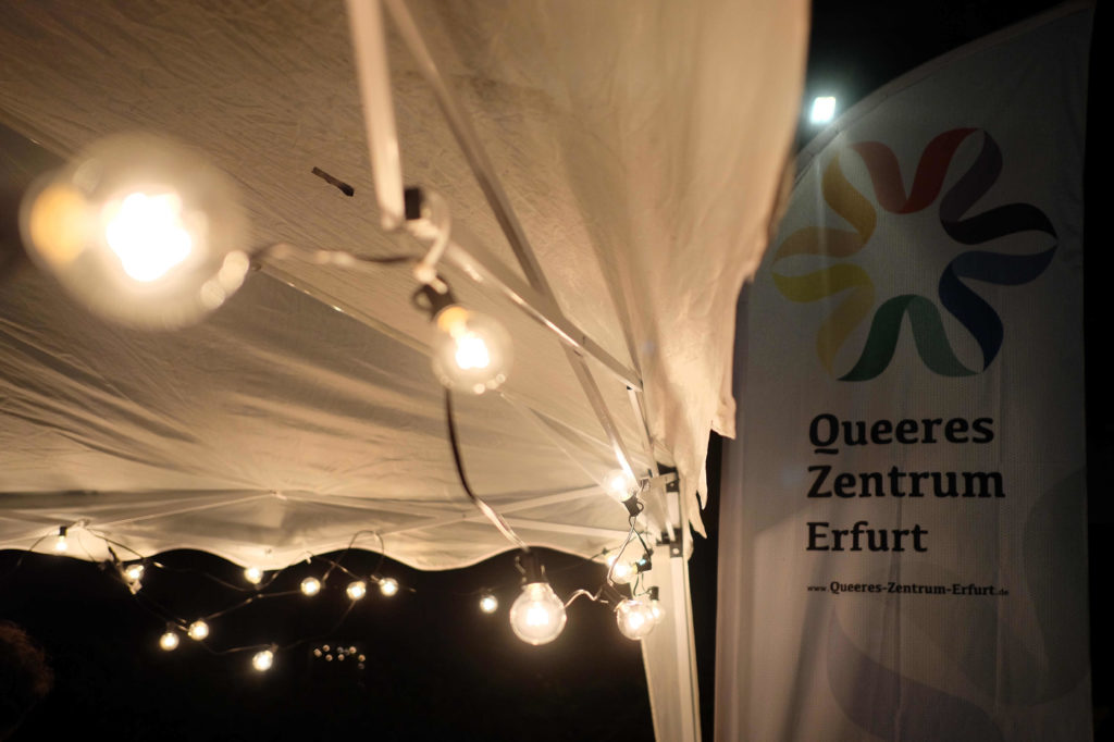 Beachflag mit Aufschrift "Queeres Zentrum Erfurt" vor weißen Pavillon mit Lichterkette